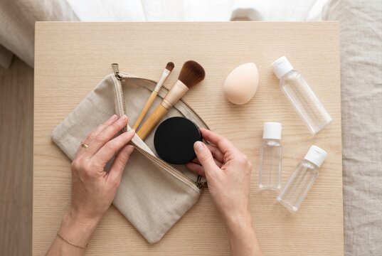 Hands packing a cosmetic compact and makeup brushes into a linen zipper pouch