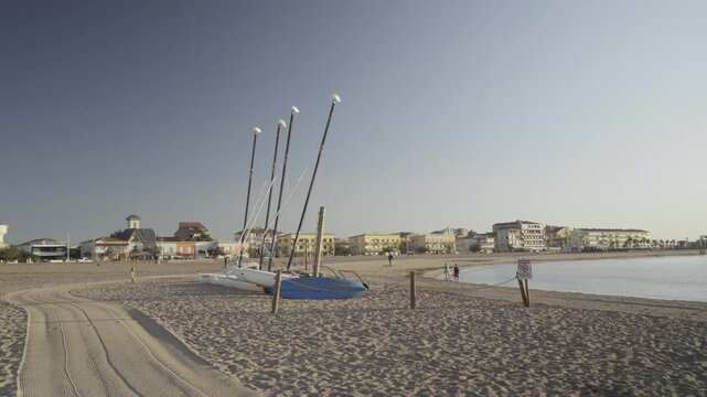  France, Valras Plage August 08, 2026 &mdash; Handheld shot of small sports catamarans parked on the sandy beach of a Mediterranean resort town with coastal buildings under a clear blue sky