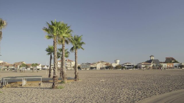 France, Valras Plage August 08, 2026 &mdash; Handheld shot of beautiful green palm trees on a wide sandy beach with coastal resort buildings and houses in the background under a clear blue sky