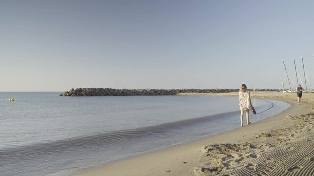 France, Valras Plage August 08, 2026 &mdash; Handheld shot of a woman in a summer dress standing on a sandy beach looking at a calm sea bay with a stone breakwater pier on the horizon during sunrise
