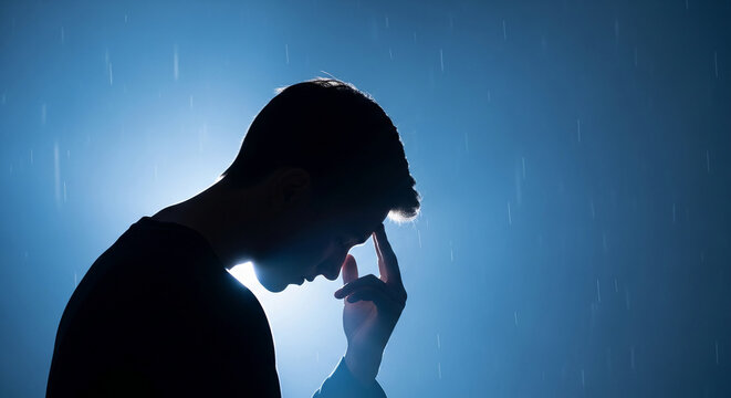 Silhouette of a man with his head bowed and hand on forehead against a blue, rainy background, symbolizing contemplation, stress, or deep thought