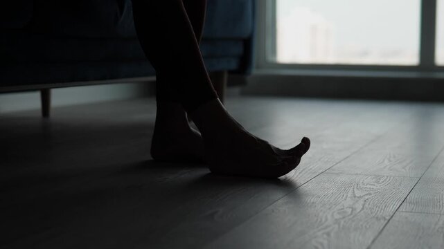 Low-angle view shows silhouetted lower legs and bare feet beside upholstered chair, with one foot gently shifting above light wood floor in bright window backlight, calm minimalist setting