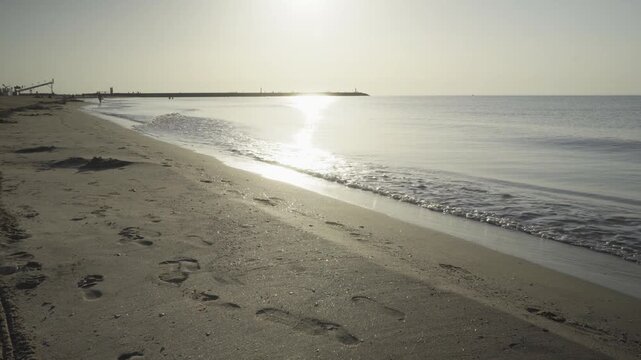 France, Valras Plage August 08, 2026 &mdash; Handheld shot of human footprints on a sandy beach shoreline with a bright golden sunrise reflecting on calm sea water and a long pier silhouette on horizon
