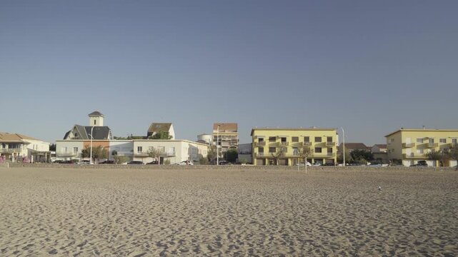 France, Valras Plage August 08, 2026 &mdash; Handheld shot of a wide empty sandy beach with a row of coastal resort town buildings, hotels and a small church under a clear blue sky on a bright sunny day

