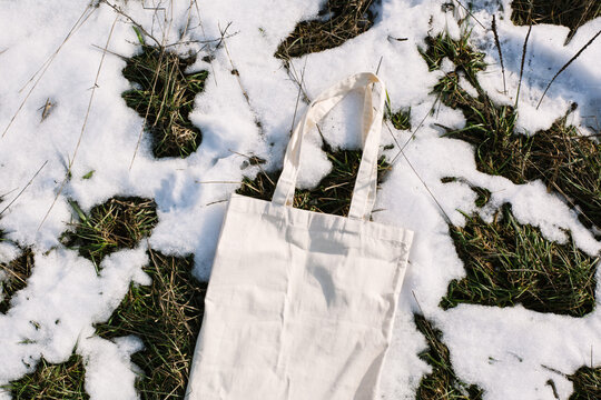 White cotton mesh bag on spring snow, green grass thawed patches background. Zero waste, no plastic, eco friendly shopping, recycling concept. Blank mockup shopper, place for text. copy space flat lay