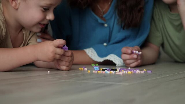 Close low angle indoor family moment with child smiling beside two adults, all handling tiny colorful blocks on wooden floor, showing careful play, learning, bonding, and shared attention