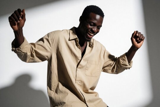 Joyful Black man in beige shirt dances with energy in studio. Happy male person shows pure excitement, freedom through movement. Cheerful guy laughs jumping moving to rhythm against white background.