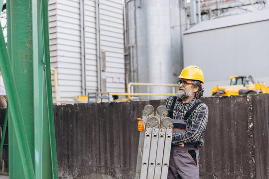 Senior worker wearing hard hat and safety glasses carrying a ladder at a large industrial granary elevator