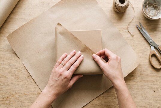 Hands wrapping a square cardboard gift box in plain brown kraft paper on a wooden desk