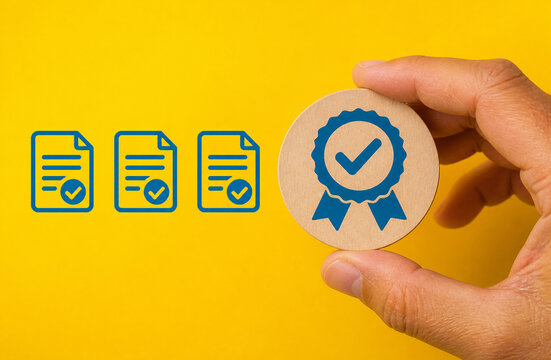 Hand holding wooden block with blue quality certificate badge next to document icons, standard compliance, warranty badge, quality assurance concept stock photo