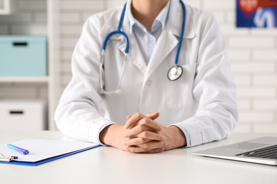 Doctor in white coat with stethoscope and clasped hands sitting at a desk with a laptop and clipboard, the background is a blurred office interior with shelves and a white brick wall