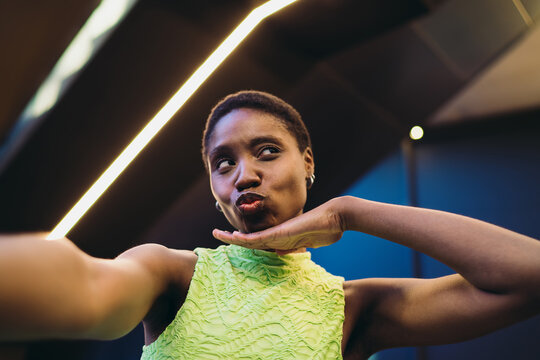Joyful African American woman sending air kiss or making duck face during smartphone selfie capture. Cheerful dark skinned model posing for social network publication near neon illumination.