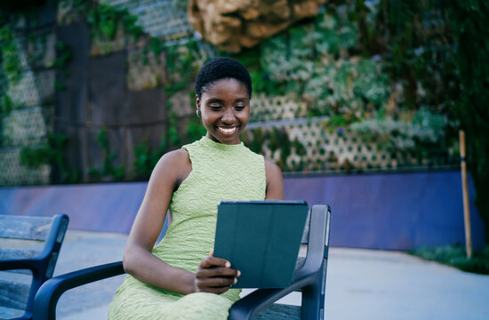 Smiling African American woman sitting on park bench and checking tablet screen. Happy dark skinned female student scrolling social media feed or reading notifications outdoors.