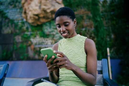 Smiling African American woman sitting on park bench and texting on green smartphone. Happy dark skinned female student checking notifications or scrolling social media feed outdoors.