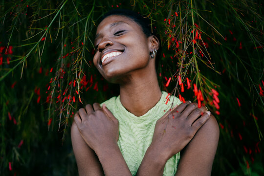 Overjoyed African American woman hugging herself with closed eyes against green leaves backdrop. Sincere dark skinned female youth experiencing self love and mindfulness in park.