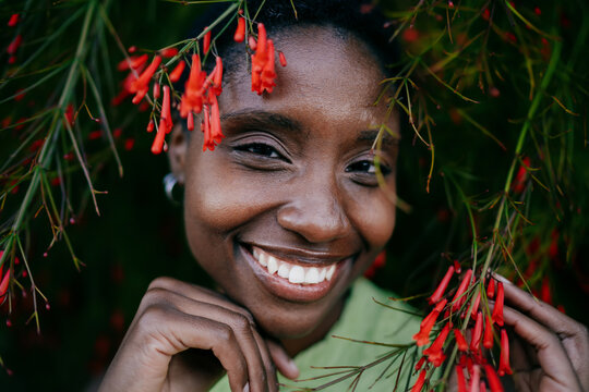 Extreme close up of happy African American woman face framed by green leaves and red flowers. Joyful dark skinned female youth looking at camera with radiant toothy smile.