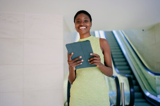 Portrait of cheerful black female student holding tablet computer on modern terminal background. Smiling African American youth with short hair checking online banking application near escalator.