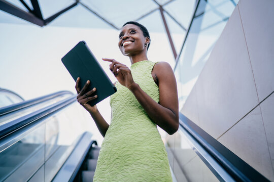 Smiling african american woman standing on city escalator, holding digital tablet and looking away toward the sky. young black female is checking a local transit schedule app