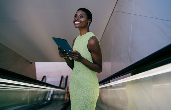 Smiling african american woman standing near an escalator handrail, looking upward with a cheerful expression while holding her tablet computer. stylish black female is wearing a lime green dress