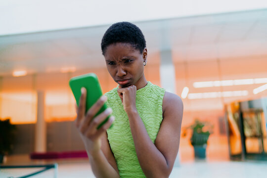 Pensive African American woman student thinking about mobile phone message. Thoughtful dark skinned model holding smartphone near chin, looking down with serious emotion inside terminal lobby.