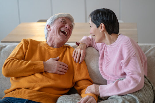 Funny cheerful senior husband and wife joking and sharing stories while sitting on couch together, happy elderly married couple in positive mood, smiling man and woman talking and having fun at home.