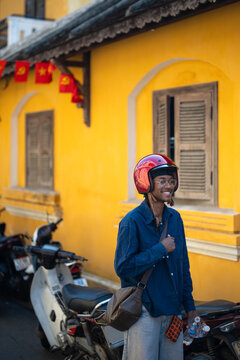 Smiling cheerful man black wearing red motorcycle helmet posing beside vintage cafe in Vietnam, happy african guy in stylish outfit and helmet standing on street outside old restaurant near motorcycle