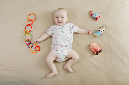 Cute 7 month baby boy lying among various playful colorful toys on beige blanket. Infant surrounded by vivid objects participating actively in sensory exploration and learning. 
