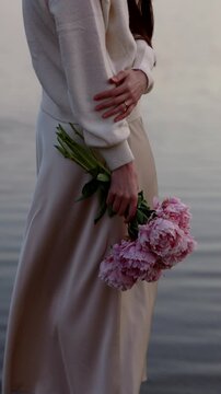 Faceless woman posing with pink peonies by water, vertical video