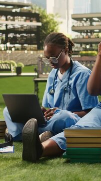 Vertical frame: Medical school peers in scrubs review laptop notes on campus, sharing ideas through tutoring, teamwork and outdoor group study for healthcare training.