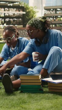 Vertical aspect: Medical students smile and exchange ideas with a laptop and coursework notes during a relaxed outdoor group study session on a modern university campus.