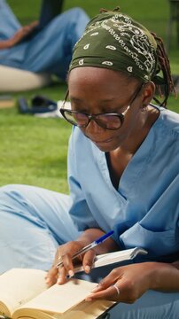 Vertical screen: The focused medical student in blue scrubs writes in an open textbook on grass at a modern campus, sitting with stacked books and study material during an outdoor learning session.