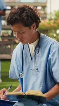 Vertical frame: a medical student reading an open textbook outdoors in blue scrubs with a stethoscope, studying beside stacked books during focused learning on a modern university campus.