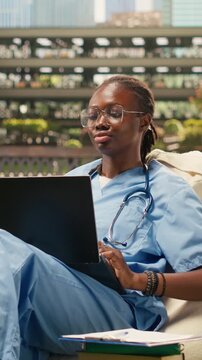 Vertical video: a Black medical student smiling and typing on a laptop while seated on a beanbag in a bright modern campus courtyard during outdoor medical school training, with study notes nearby.