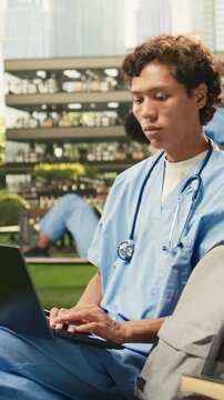 Vertical aspect: The male medical student studies independently on a laptop while seated on a wooden bench in a modern campus courtyard, representing international medical education and student life.