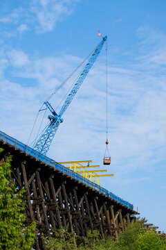 Old wooden railroad train trestle construction project, blue crane holding work basket over job site, maintenance on structural framework, sunny summer day, United States
