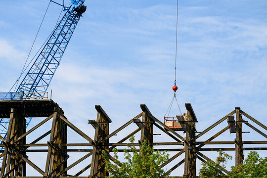 Workman in safety equipment in work basket help aloft by crane, old wood railway train trestle construction project to update structural framework, sunny summer transportation project, United States
