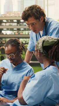 Vertical frame: The group of peers in blue scrubs discusses coursework on a campus bench, using a laptop, open book and materials for outdoor learning, student collaboration and medical training.
