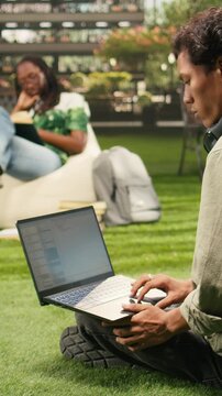 Vertical video: The international student uses a laptop with coding content on a campus bench as classmates study in the background for outdoor coding and automation study in an academic environment.