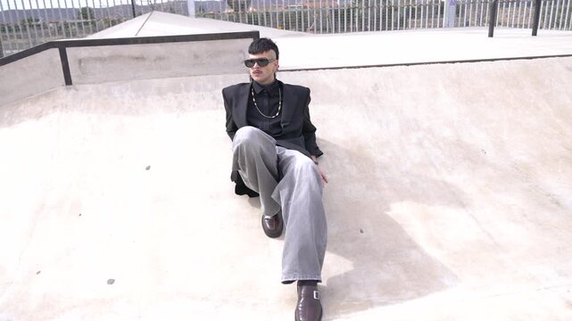 A full-body shot of a young man wearing a stylish outfit consisting of a black jacket, sunglasses, earrings, and piercings, looking at the camera and to the sides while sitting on an outdoor skate par