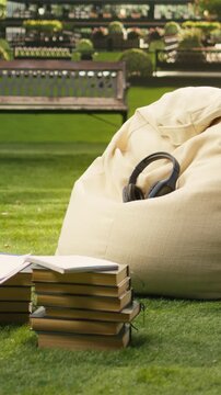 Portrait format: Wireless headphones rest on a fabric beanbag outdoors, framed as technology detail with stacked books, greenery and modern buildings on campus blurred behind, reflecting student life.