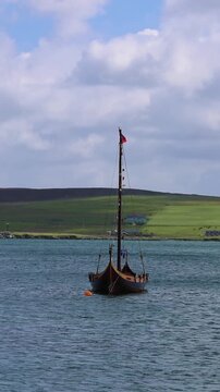 Traditional wooden Viking ship floating in clear bay waters near Lerwick Shetland Islands with green rolling hills behind