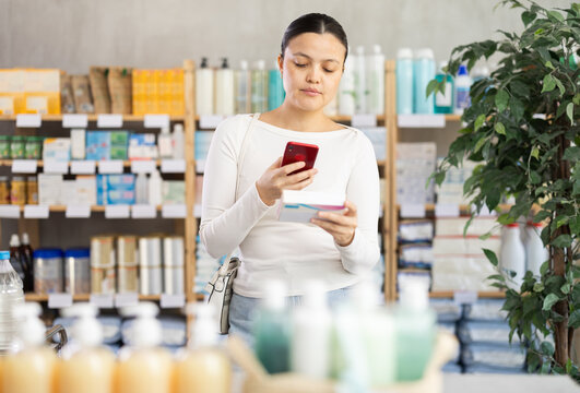 Asian female shopper in pharmacy scanning barcode on box of pills with smartphone, carefully checking dosage, side effects, or contraindications before purchase, ensuring safe self-medication