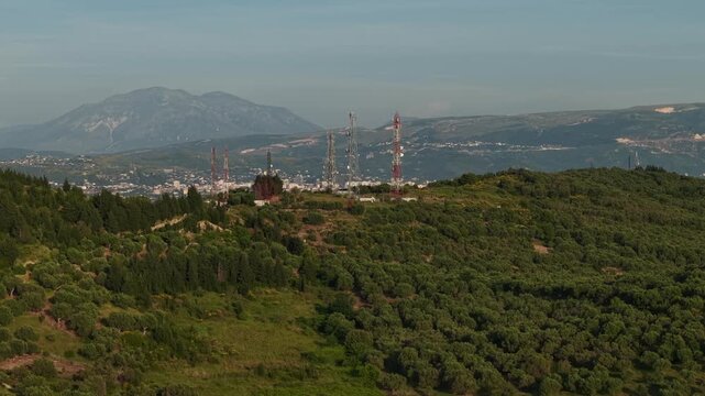 Aerial view of communication towers on a green hill overlooking the town with mountains in the distance in Zvernec, Vlore County, Albania.