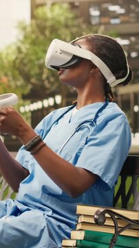 Vertical video: The woman medical student wears VR glasses and raises the controllers during medical training on campus, sitting on a bench in blue scrubs, using VR for healthcare learning.