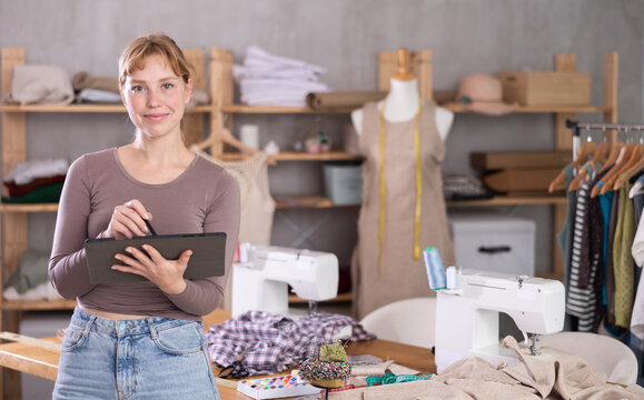 Qualified seamstress standing in front of sewing workshop, worktable and mannequin with tablet. Fashion designer accepts online orders for custom tailoring