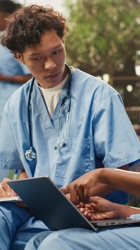 Vertical video: Medical students discuss coursework with a laptop and study materials, sitting on a bench near books on a campus, with classmates in a group study session, during peer tutoring.