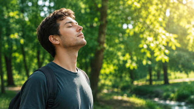 A young man with closed eyes enjoys fresh air in a sunlit forest, surrounded by lush green trees and dappled sunlight.