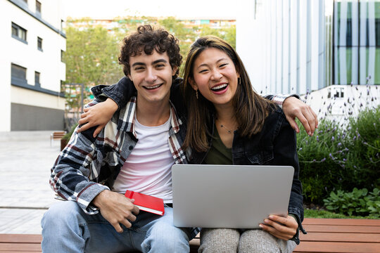 Cheerful students using laptop and smiling outdoors in university campus looking at camera smiling