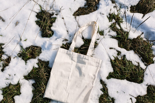 White cotton mesh bag on spring snow, green grass thawed patches background. Zero waste, no plastic, eco friendly shopping, recycling concept. Blank mockup shopper, place for text. copy space flat lay