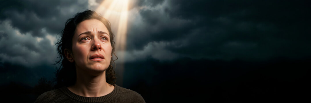 Crying woman looking up in prayer during grief. Sad female face with tears illuminated by divine light beam. Panoramic banner with copy space for faith and hope concept